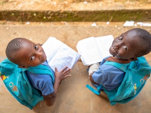Two girls, 7- and 8-years-old, from Central African Republic smile up at the camera from where they're sitting outside, wearing matching 'Education cannot wait' backpacks and holding open books.