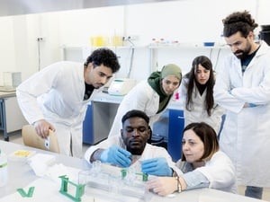 A group of people wearing lab coats gather round as a student and professor carry out an experiement in a laboratory 