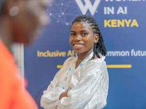 A smiling young woman with crossed arms stands in front of a conference poster for an event on women in AI in Kenya