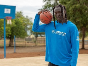 A South Sudanese man holds a basketball on his shoulder, a basketball court behind him