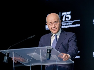 A man in a suit delivers a speech standing at a clear lectern in front of a dark background.
