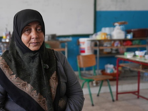 A woman in a headscarf sits in a school classroom with household items on desks in the background