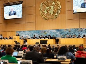 A man speaks at a lectern on a auditorium stage with people behind him seated beneath large screens while an audience looks on