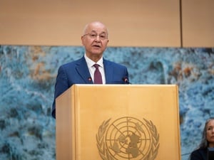 A man in a suit and tie speaks from behind a wooden lectern emblazoned with the UN logo