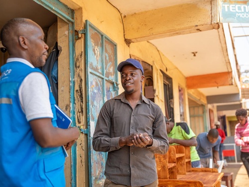Two men, one in a UNHCR vest and one without, stand talking outside a row of buildings