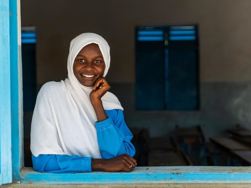A young girl smiles at the camera, looking through an open window.