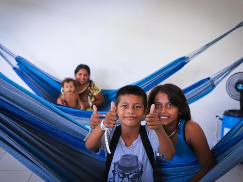 Two children sit in a hammock giving a thumbs up, with a mother and baby in the hammock behind them