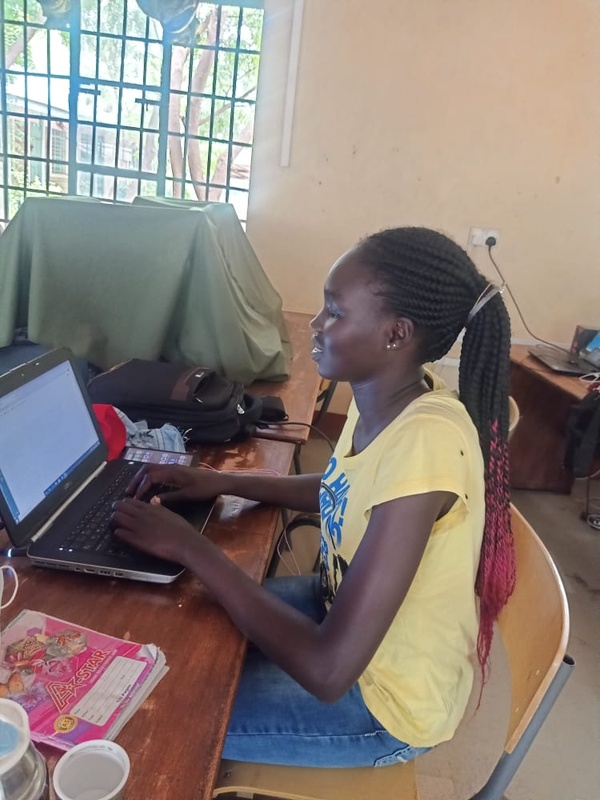 A young woman in a yellow shirt sits at a desk with a laptop in front of her