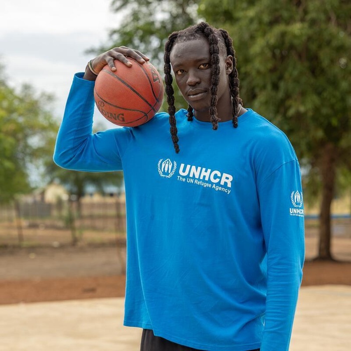 A South Sudanese man holds a basketball on his shoulder, a basketball court behind him