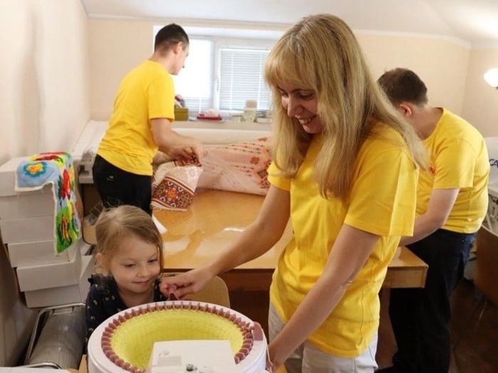 A woman wearing a bright yellow t-shirt smiles as she shows a young child a toy.