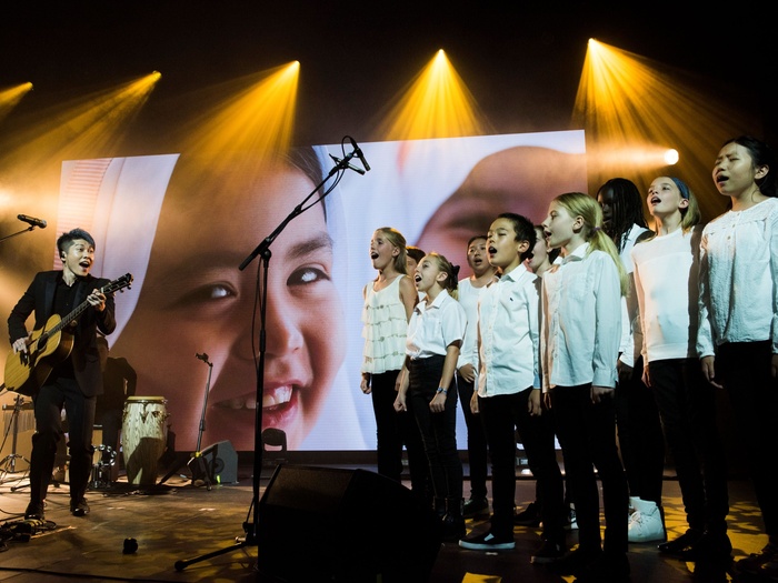 MIYAVI on stage performing with his guitar, a choir of children in white shirts and black pants to his right.