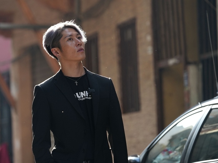 A portrait of MIYAVI wearing a black UNHCR t-shirt and black blazer, outside, staring into the distance.