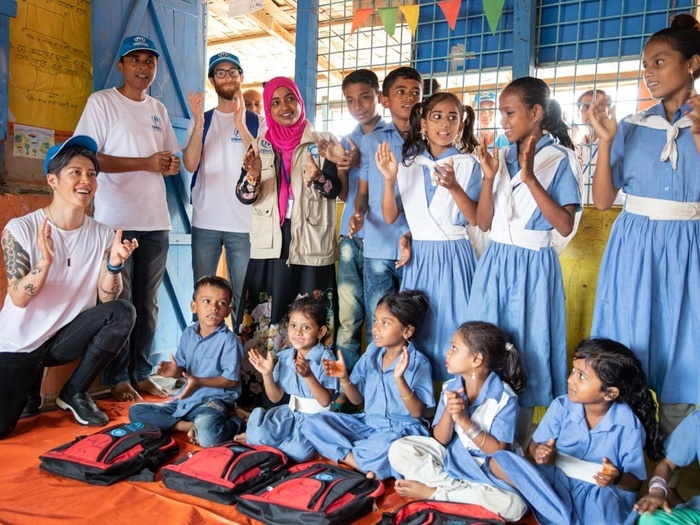 A group of students and three UNHCR staff clap along with MIYAVI, who is kneeling on the floor beside them.