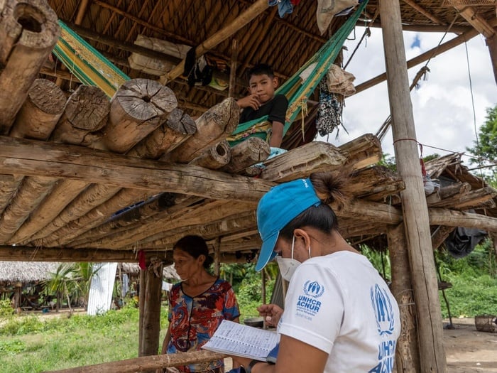 A UNHCR staff member writes on a notebook in front of a woman, and a young boy sitting on a hammock.