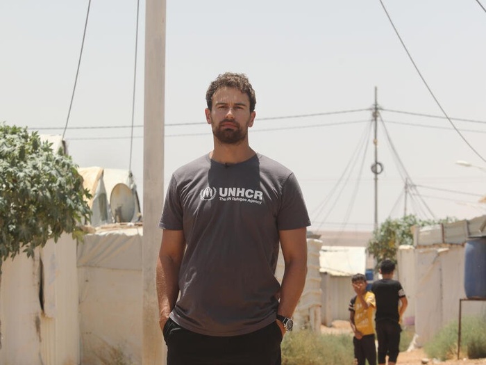 A man stands in the middle of a refugee camp, wearing a t-shirt with the UNHCR logo.