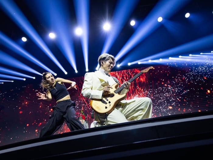 MIYAVI on stage on his knees playing a guitar, a dancer and bright lights behind him