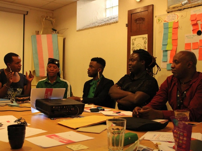 A group of people sit inside, around a table with papers, glasses, laptops and an overhead projector on it. There is a trans flag in the background and a whiteboard with post-its.