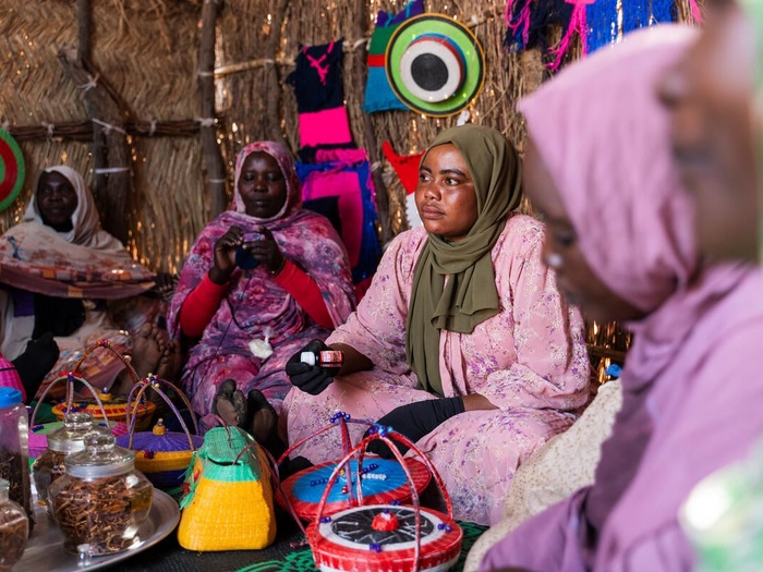 Women sit on the floor of a shelter showing off crafts they have made.