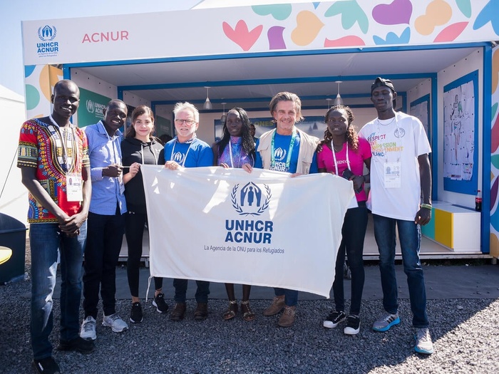 Two UNHCR representatives and members of the Refugee Olympic Team hold up a UNHCR/ACNUR banner