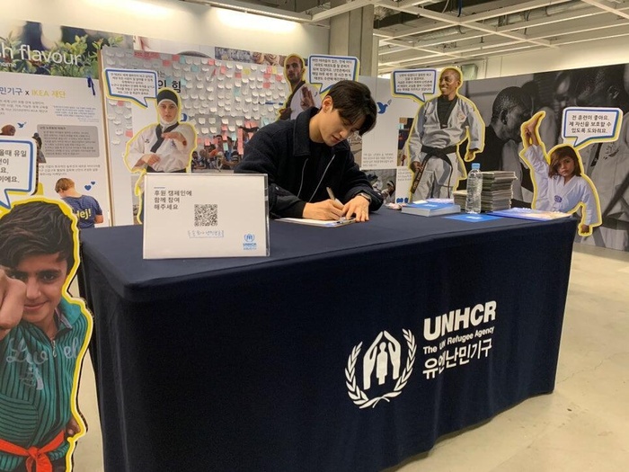 Daehoon Lee signs an autograph sitting at a table with UNHCR Korea branding. Surrounding him are cut-out figures of taekwondo poses.