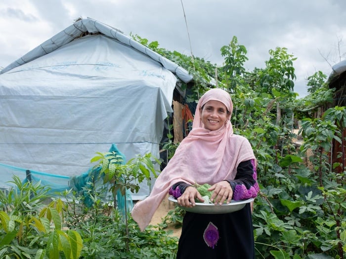 A woman holding a plate of fresh vegetables while standing in front of a garden in a refugee camp. 