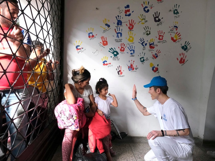 MIYAVI high-fives a young child with a wall of painted hand prints behind them. Others stand nearby behind a fence.