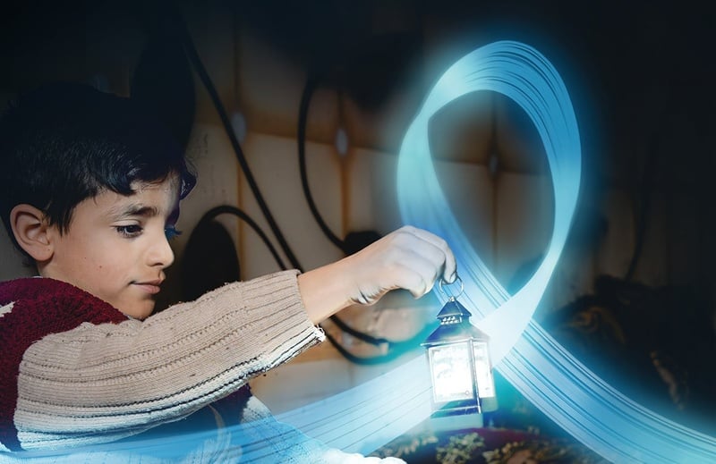 A young boy is holding a small lantern in a dark room. A blue, shiny and flying ribbon floats around the lantern.