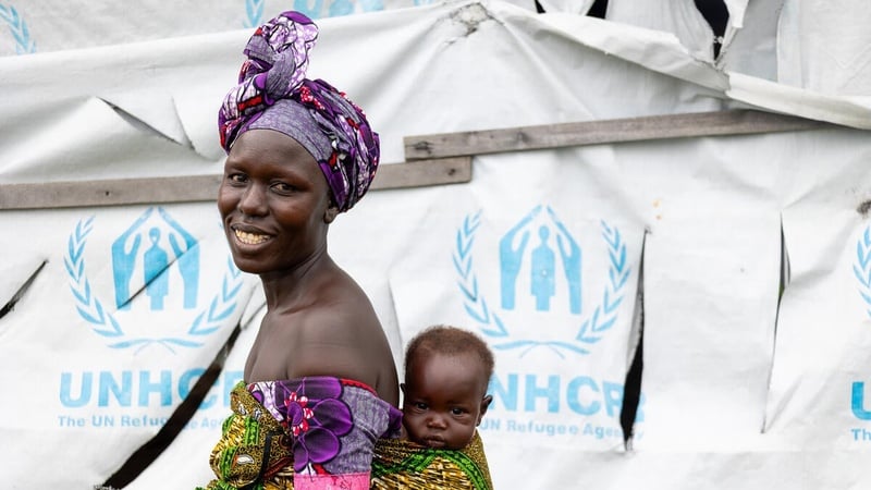 A smiling woman carries her baby boy on her back in front of a UNHCR tent.