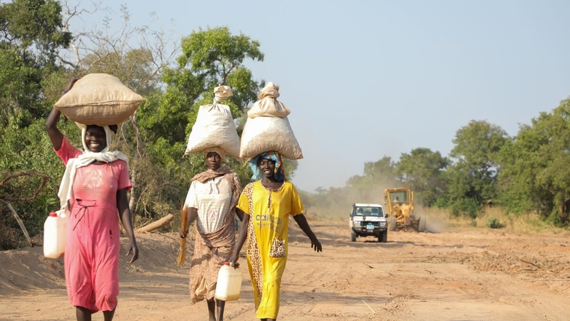 Three friends, Sudanese refugee women, walk together along a dirt road, bags balanced on their heads, smiling and laughing.