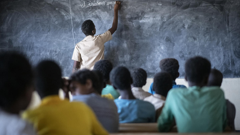 A man writes on a blackboard in a classroom of teenagers. 
