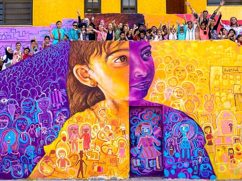 A group of girls standing over the mural they painted. The mural represents a girl, holding a tea pot.