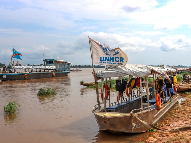 Wooden boats float on a wide, muddy river. One bears a UNHCR flag.