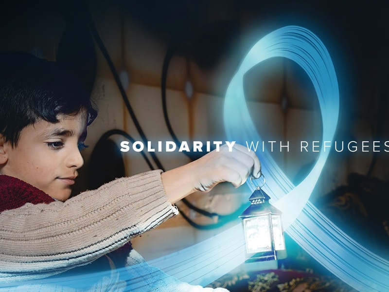 A young boy is holding a small lantern in a dark room. A blue, shiny and flying ribbon floats around the lantern. The text reads "solidarity with refugees".