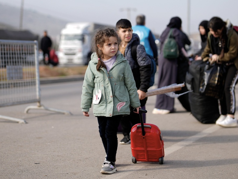 A young child walks on asphalt pulling a small suitcase.