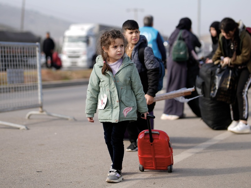 A young child walks on asphalt pulling a small suitcase.