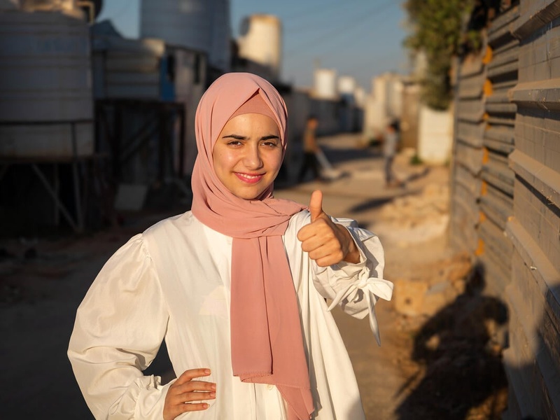 A young woman in a pink headscarf smiles confidently and gives the camera a thumbs up.