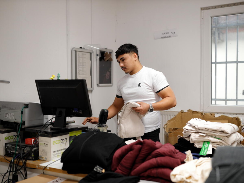 A male, 20-year-old refugee from Afghanistan stands behind a computer and piles of clothing at his workplace, a Sarajevo-based textile company in Bosnia and Herzegovina.