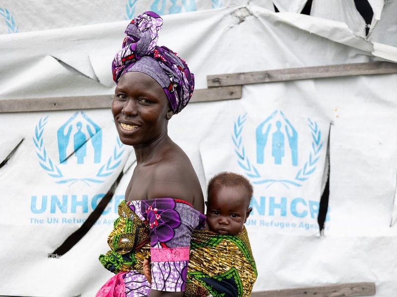 A smiling woman carries her baby boy on her back in front of a UNHCR tent.