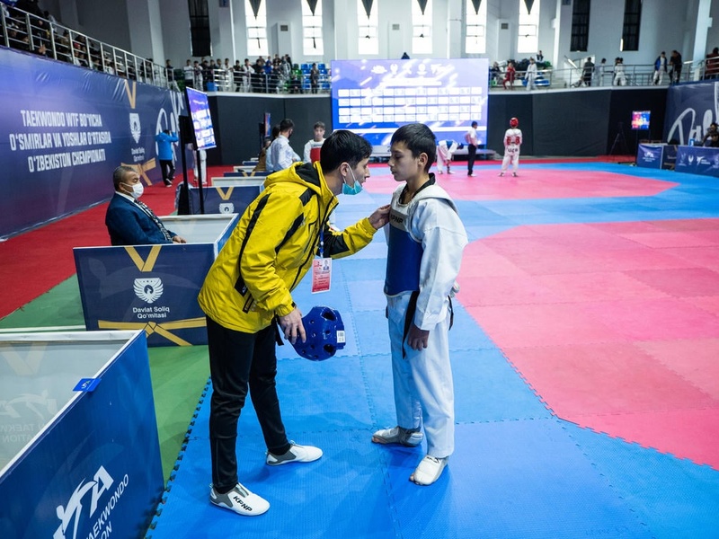 A man in a bright yellow athletics tracksuit speaks to a young child in a martial arts uniform inside a gymnasium.