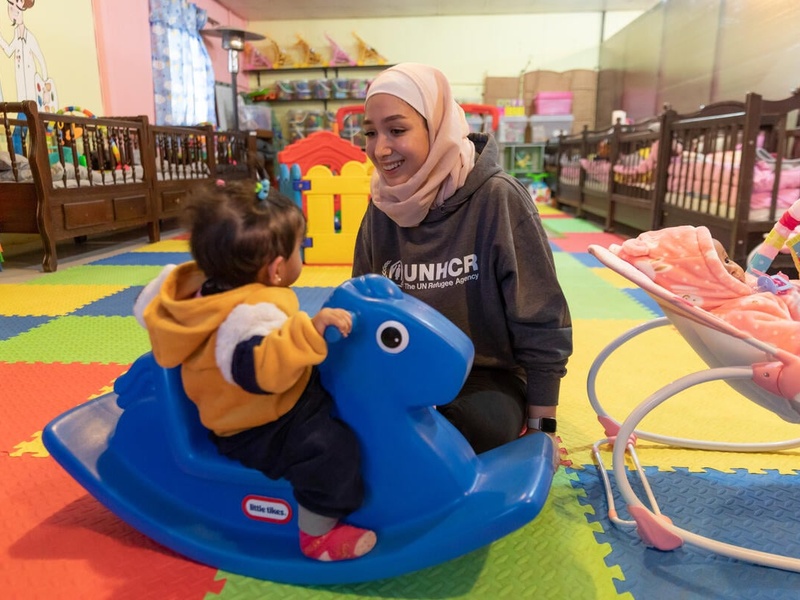 A young woman sits on the ground of a nursery and plays with a toddler.