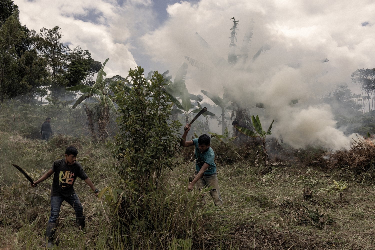 Young men clear vegetation with machetes while a small fire burns in the background