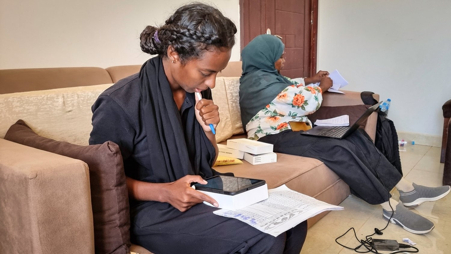 A young woman sits on a couch looking down at a tablet and a spreadsheet.