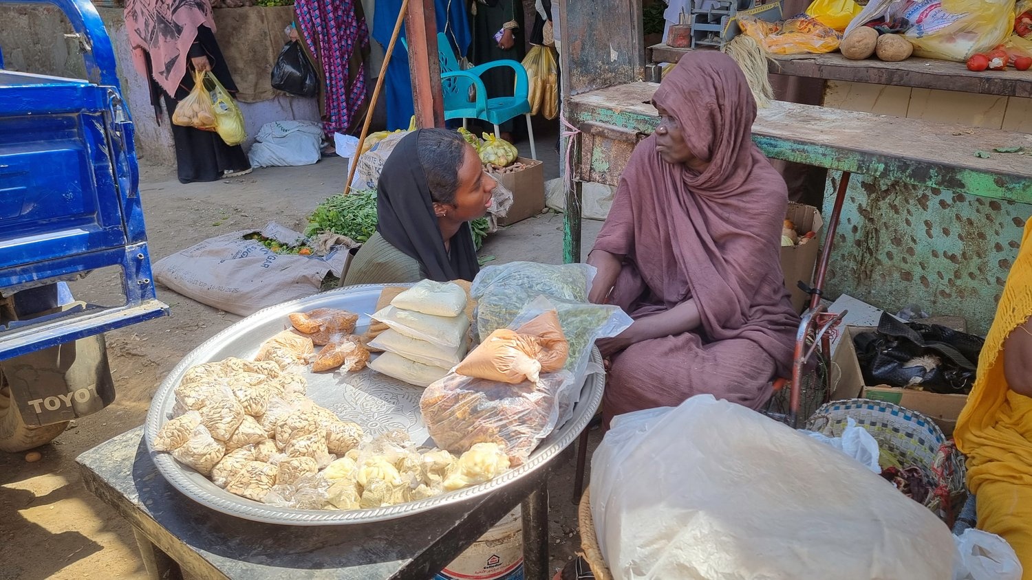 A woman crouches down to speak to a woman seated in front of a tray of food she is selling in a market.