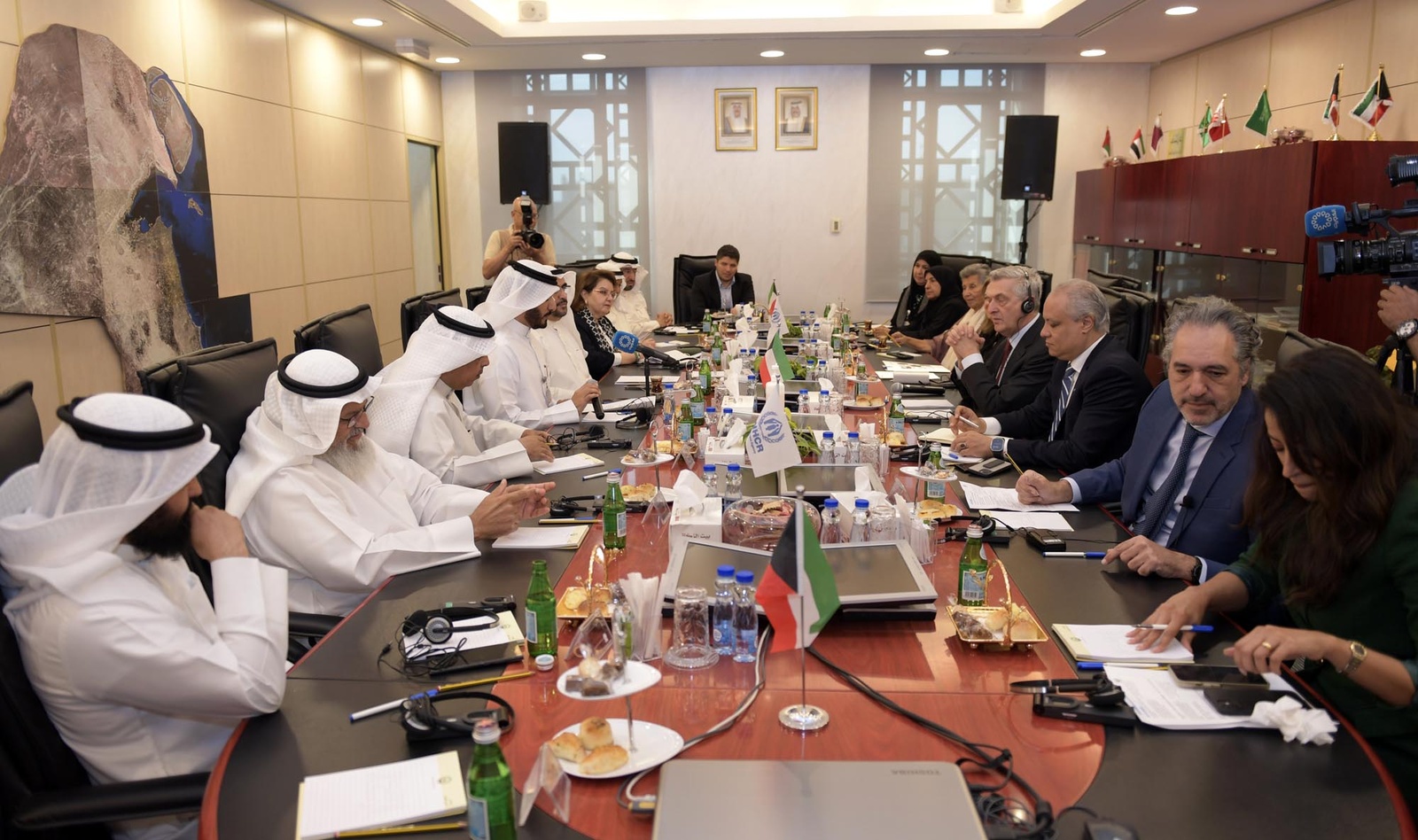 A large conference table with 15 people seated around it and a photographer in the corner