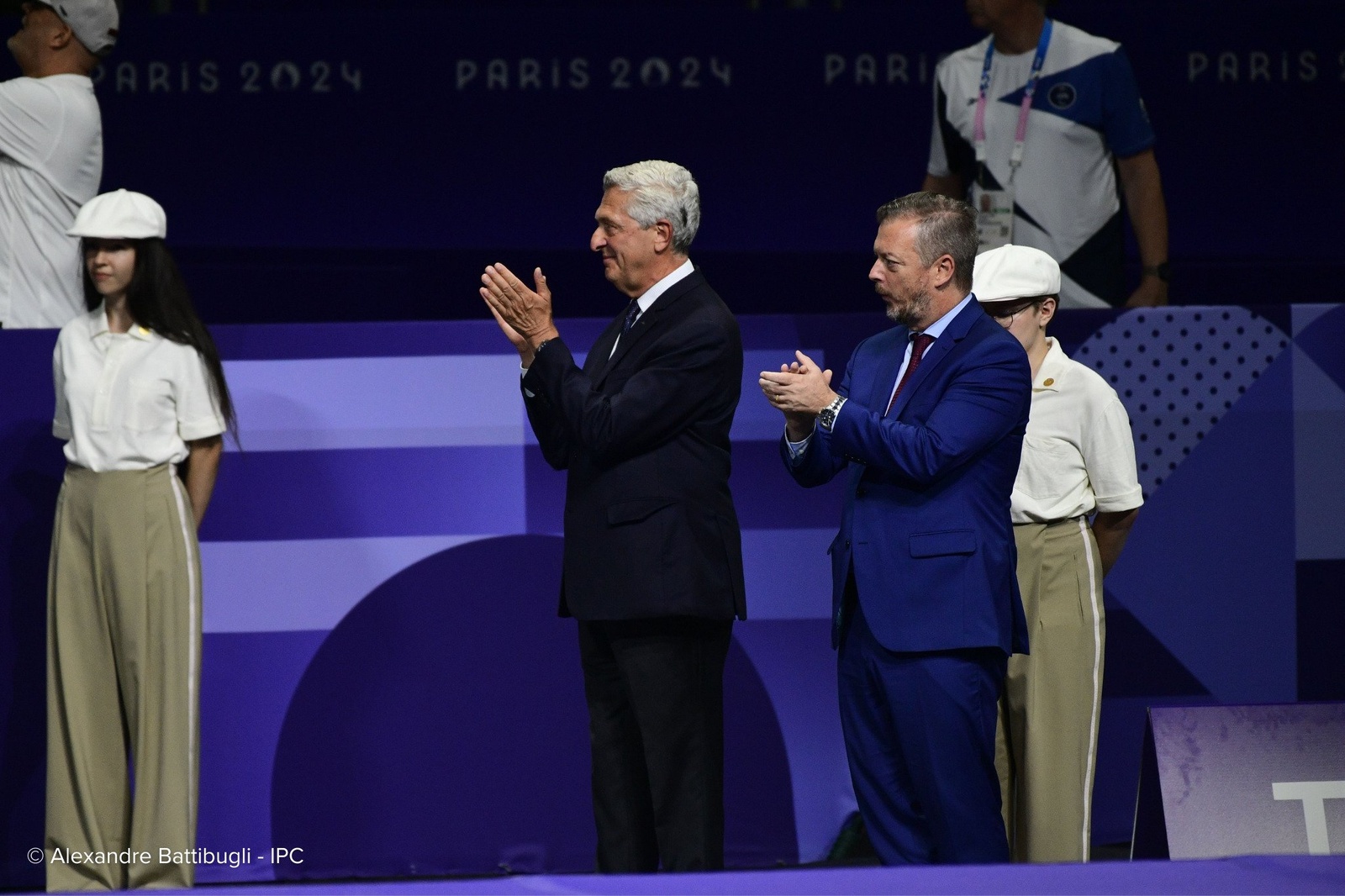 Two men wearing suits applaud at a venue displaying Paris 2024 Games logos.