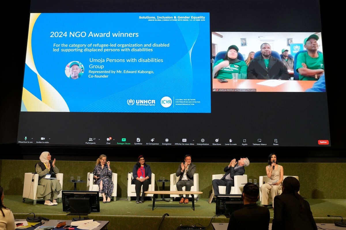 A panel of people on stage and two large screens above them, displaying a slide about the 2024 NGO Award winners and Mr. Edward Kaboyi dialling in on screen