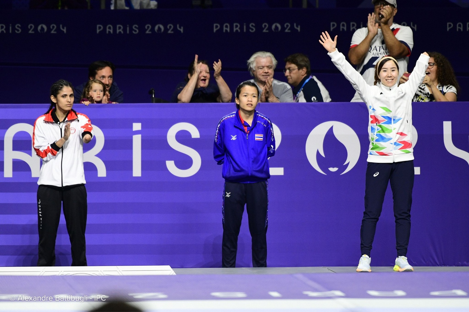 an athlete raises her arms in celebration as two others applaud while looking on.