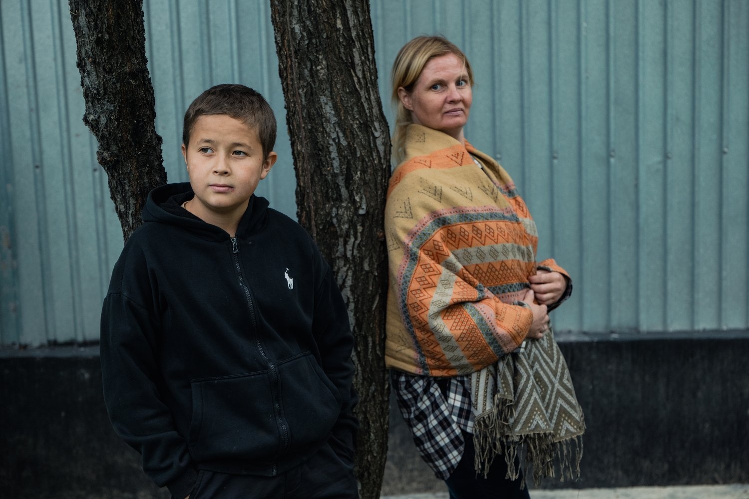 A woman wearing a shawl leans against a tree next to a boy leaning against another tree and looking away.
