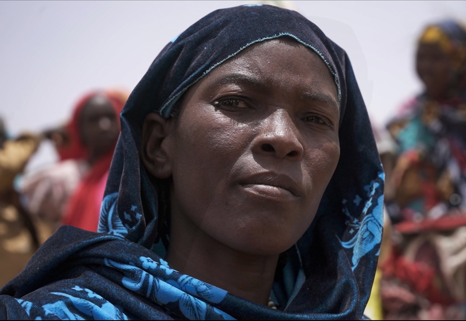 Close-up of a woman wearing a blue-patterned headscarf sitting outside with other women in the background