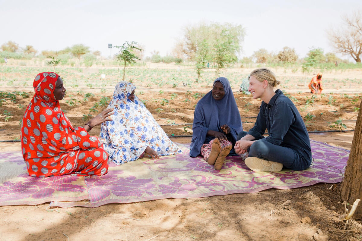 Four women laugh together sitting on a blanket in a vegetable garden.